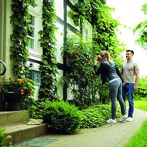 Junger Mann neben Frau, die an Rosen vor Fachwerkhaus riecht.